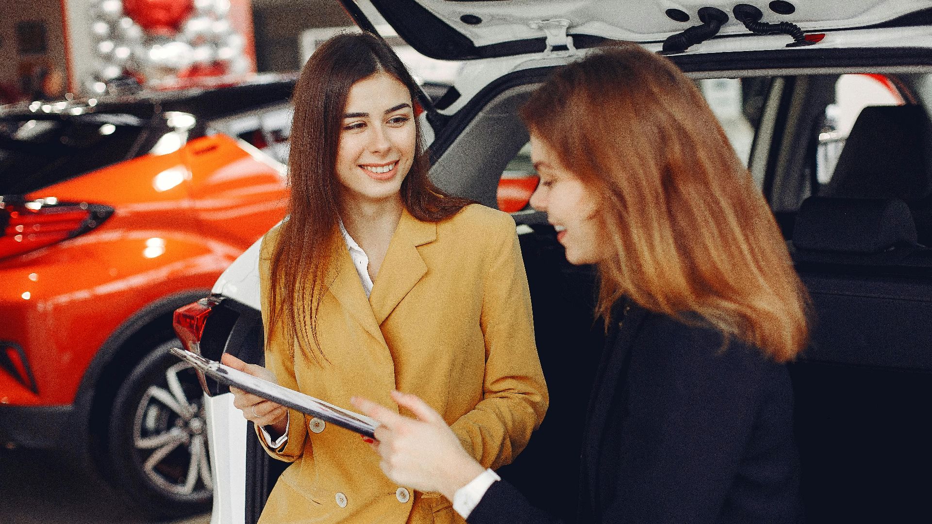Positive smiling agent in trendy formal light brown suit discussing with happy excited female customer details of contract while leaning on car in showroom
