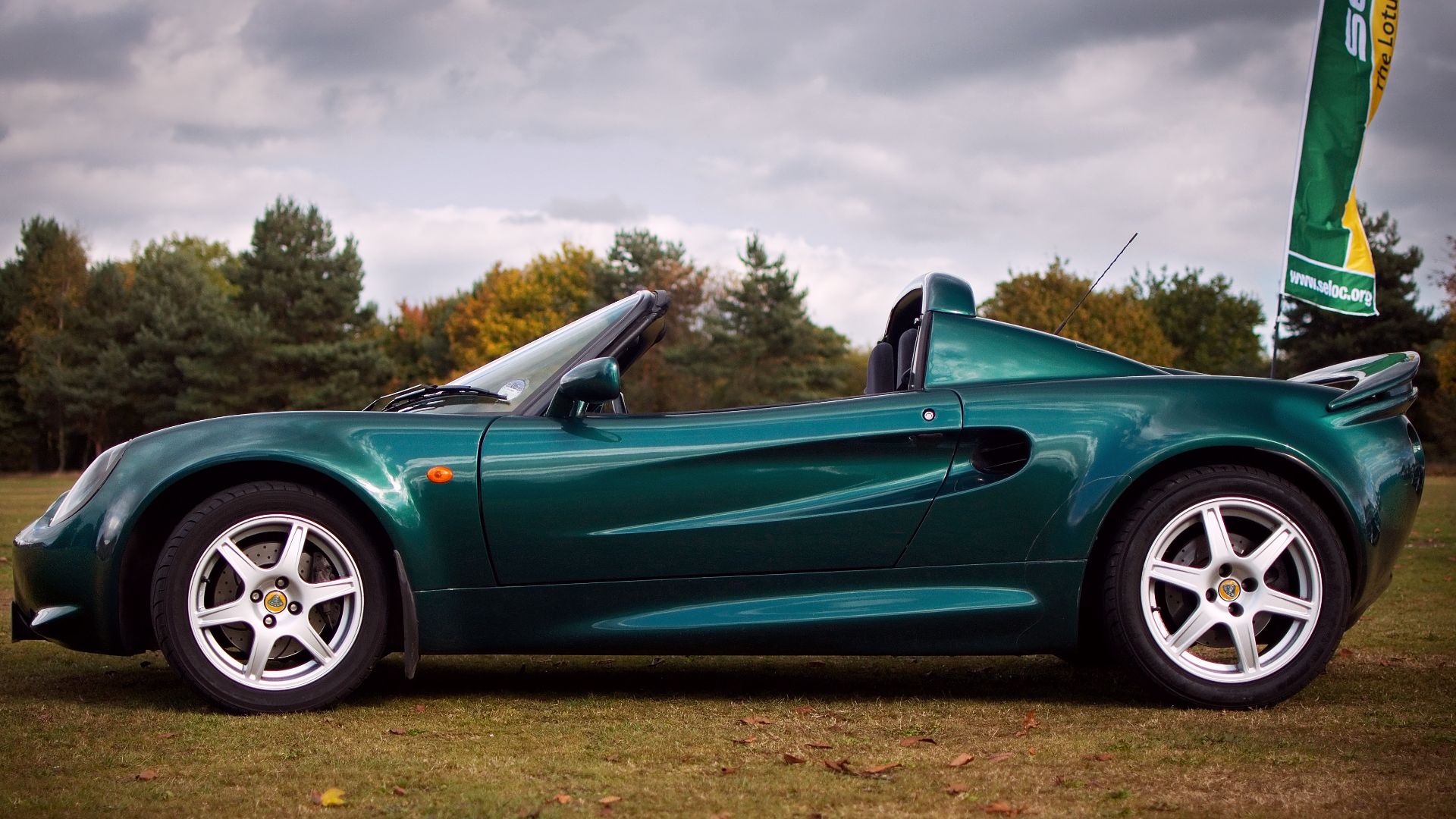 Lotus Elise Series 1, 111S version. Visual notes: as it's a 111S it has a rear spoiler, but unlike the standard 111S this one has it raised slightly on raiser blocks (like a Sport 160). The wheels are the standard 111S 6 spoke wheels. The brake discs are non-standard grooved and drilled. The paint colour is Lotus Racing Green, a metallic green.
In the background you can see a flag for the lotus owners club - I didn't deliberately park near it for the shot, I promise!