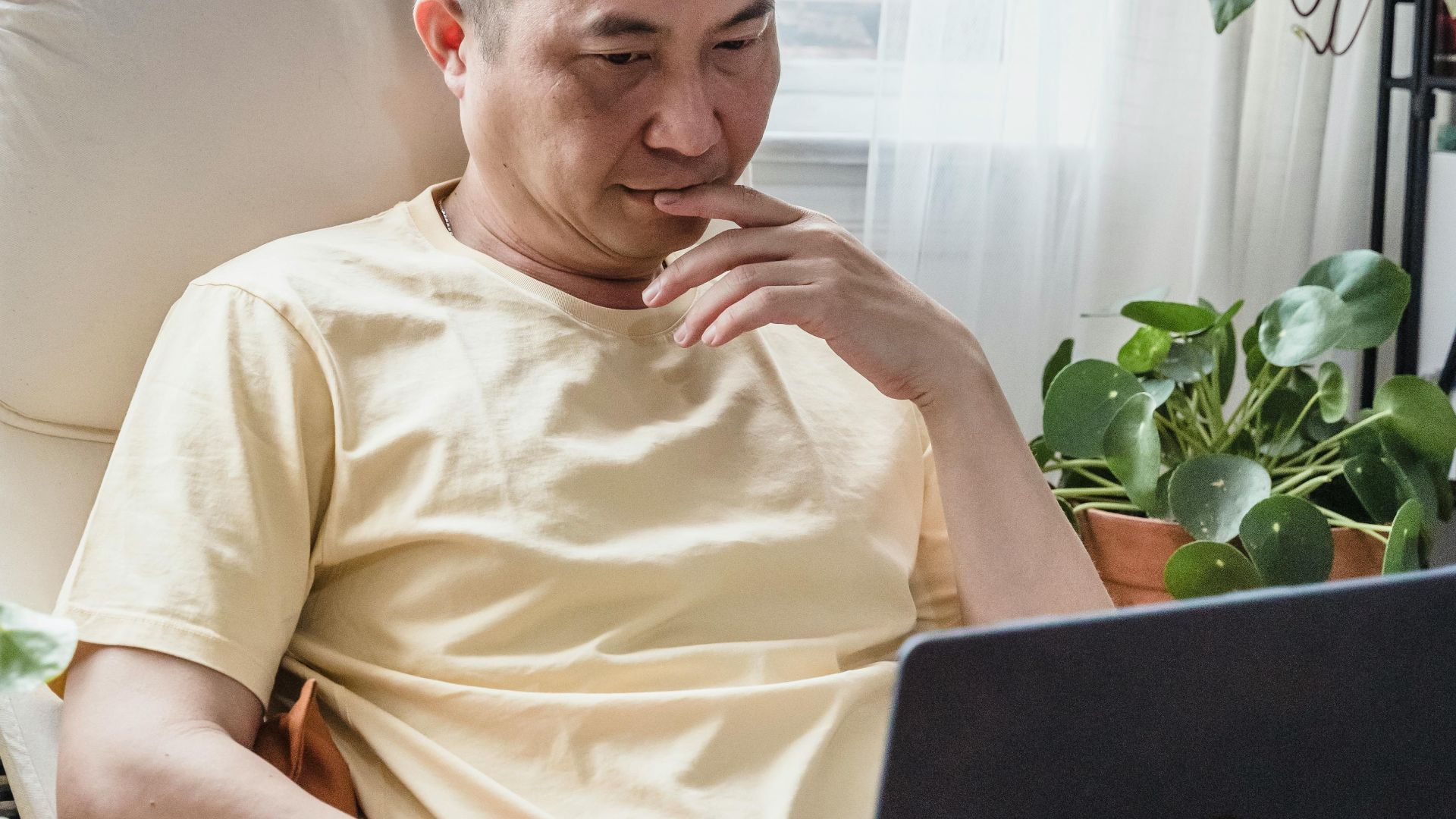 Adult sitting indoors using a laptop surrounded by plants and natural light.