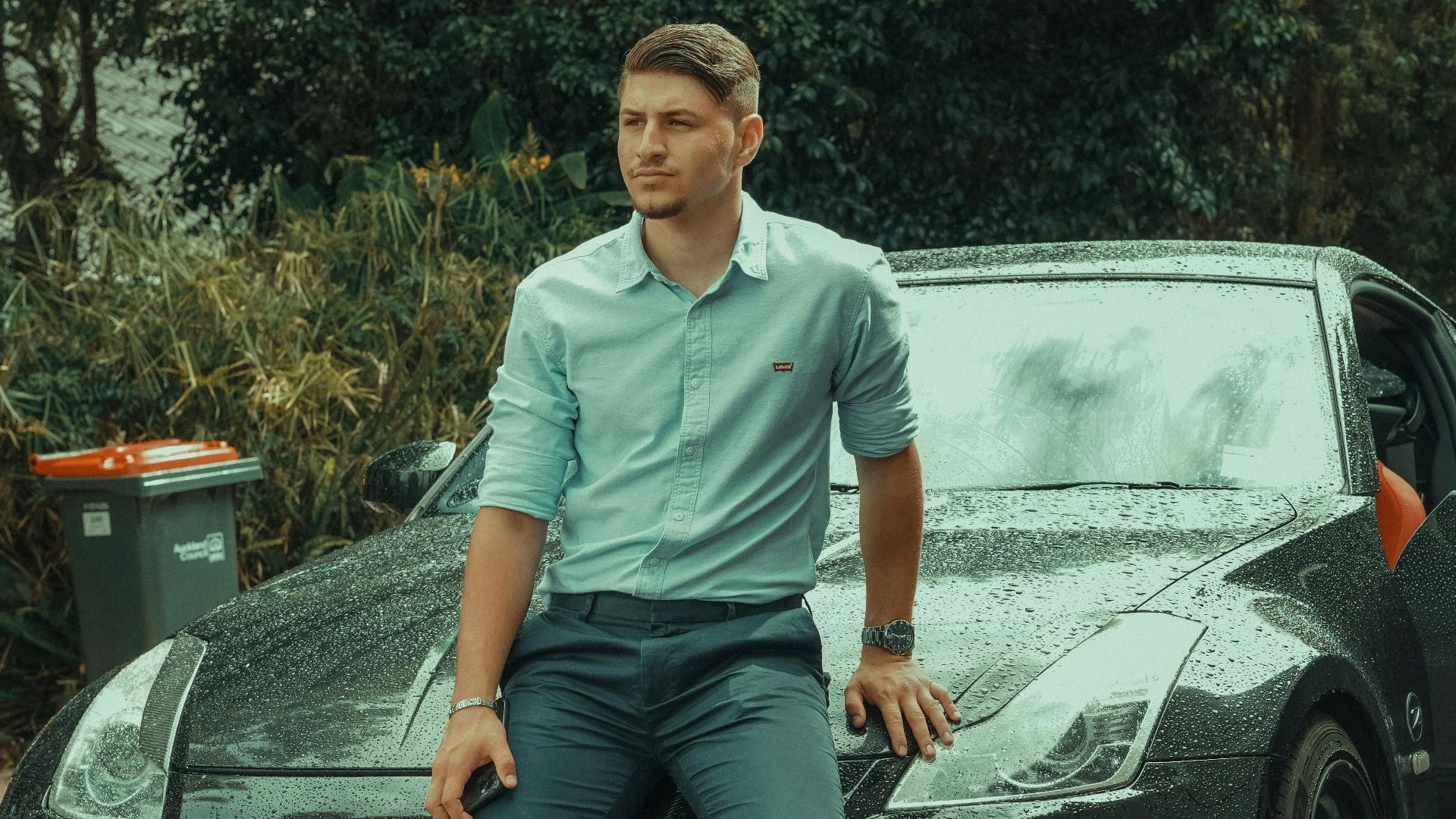 Young man in formal attire leaning on a luxury sports car parked outdoors, exuding style and confidence.