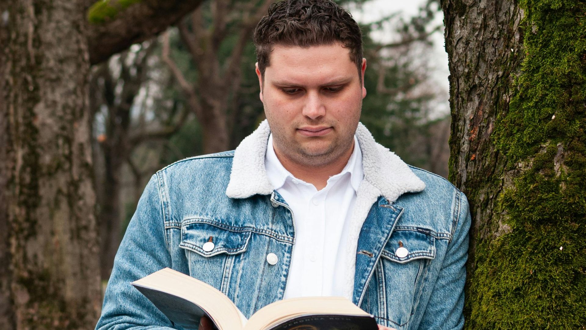 A man reads a book outdoors in Milano, Lombardia, Italia, leaning against a moss-covered tree.