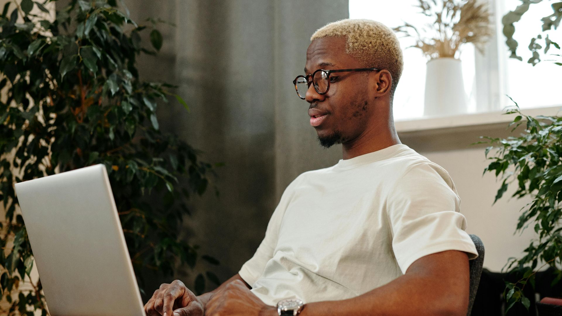 Focused young man with glasses working on a laptop in a cozy indoor space surrounded by plants.