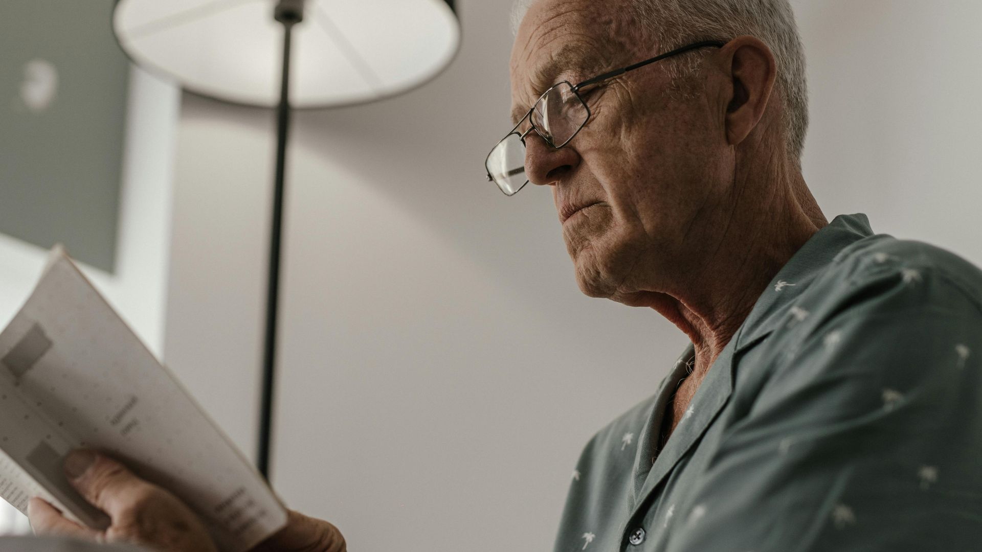 Senior man with eyeglasses reading a book under a lamp in a comfortable and serene indoor setting.