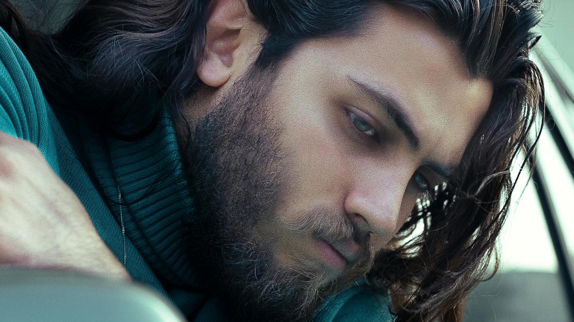 Thoughtful man in teal turtleneck looks out from car window with long hair flowing.
