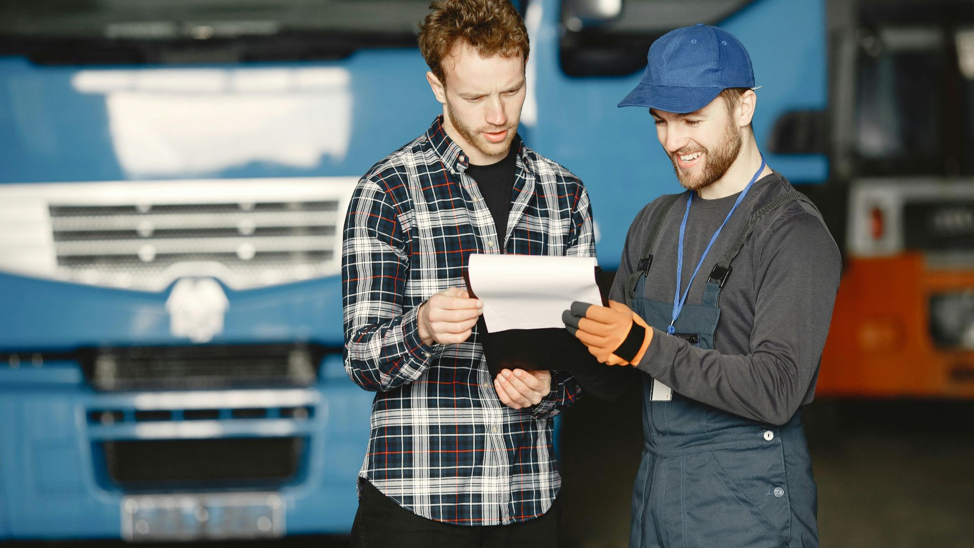 Two men in discussion over vehicle maintenance documents in a garage setting with trucks in the background.
