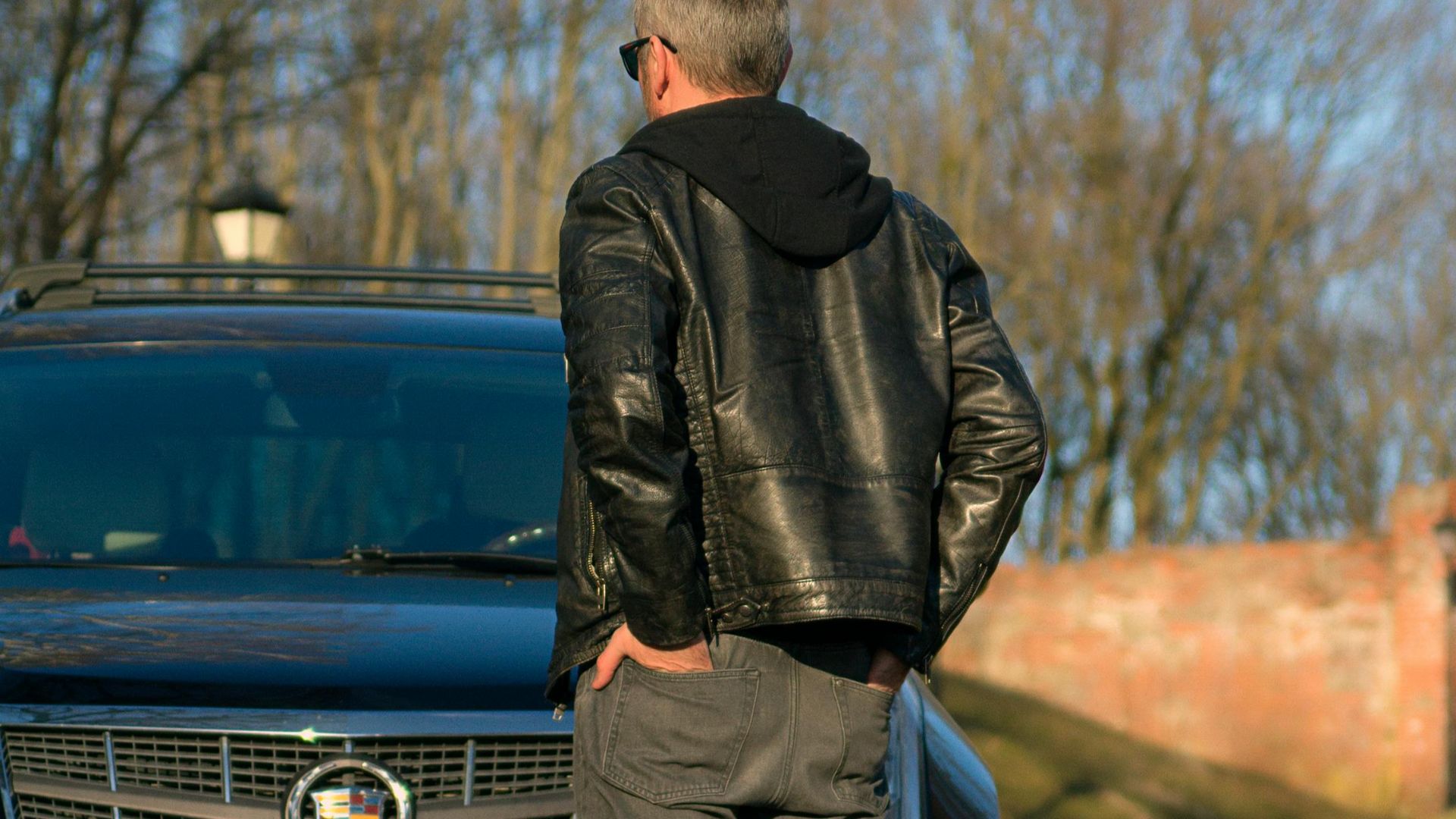 A man with hands in pockets stands by a black Cadillac outdoors.