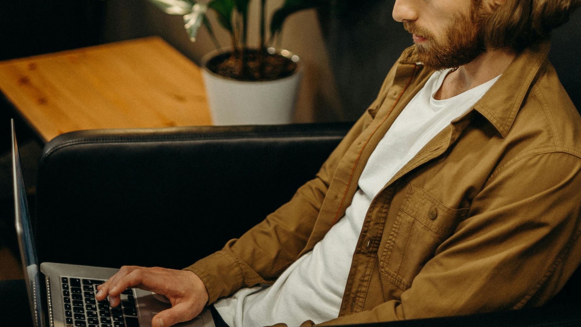 Man with laptop in cozy room focusing on remote work. Indoor setting with lamp, plant, and wall art.