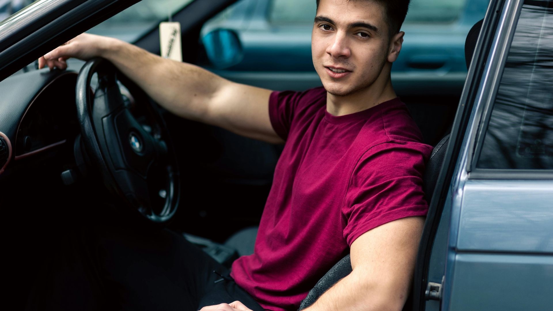 A young man wearing a maroon t-shirt sits casually in a blue car, portraying a relaxed and modern lifestyle.