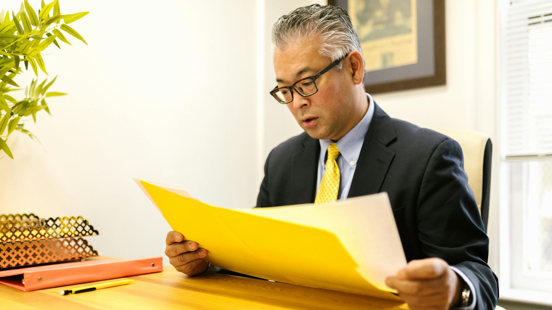 Asian businessman in corporate attire reading documents at office desk with a yellow folder.