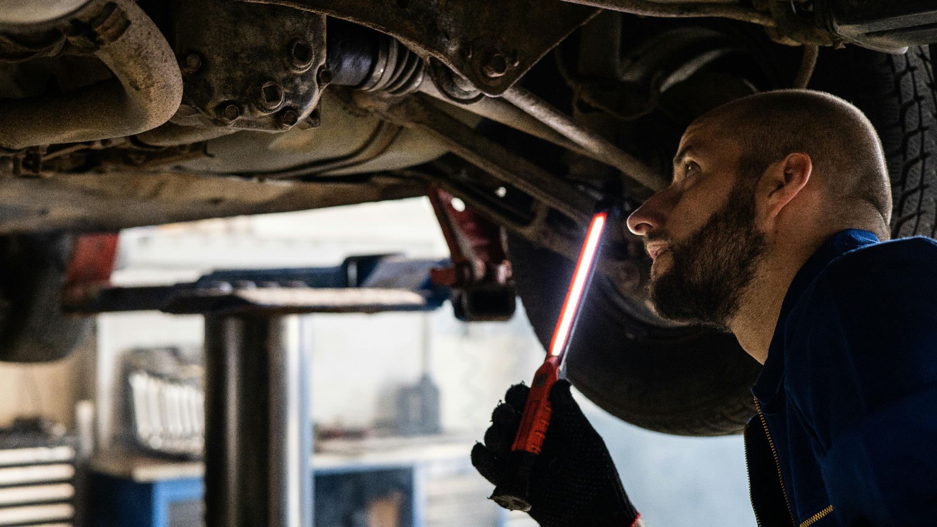 Mechanic using light to inspect vehicle undercarriage in workshop for maintenance.