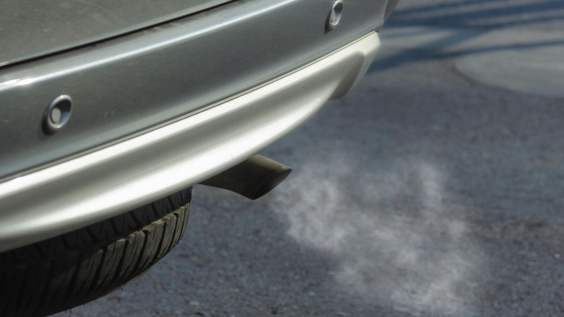 Close-up of a silver car exhaust pipe emitting smoke on asphalt road.