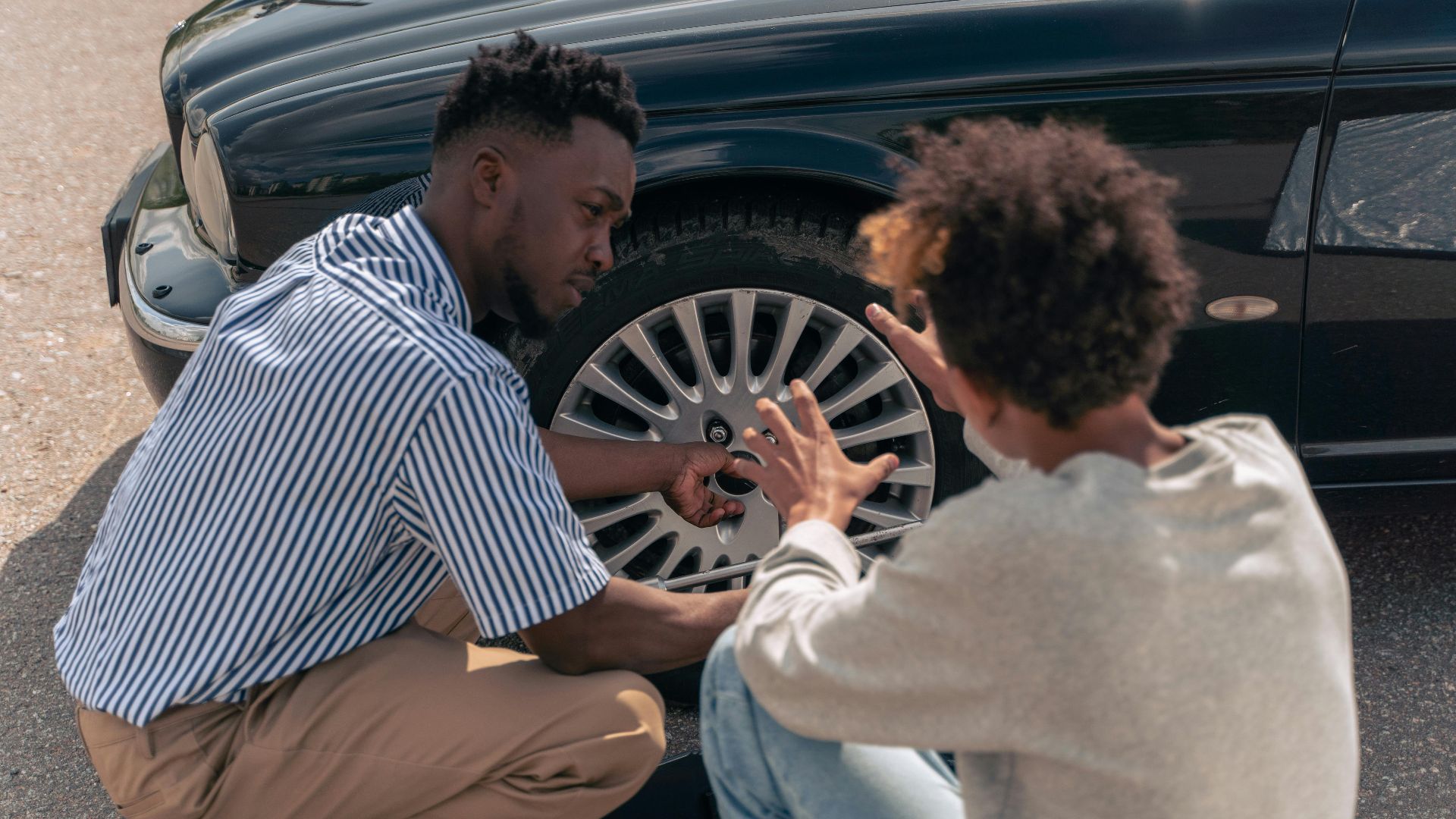A father instructs his teenage son on how to change a car tire outdoors.