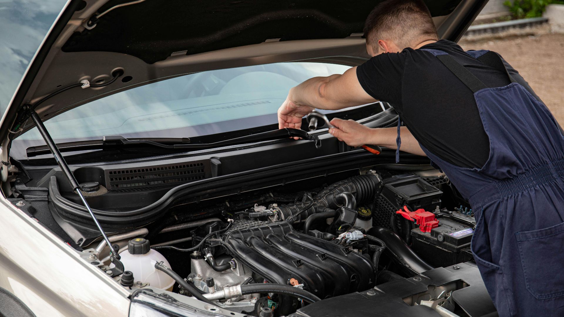 A mechanic uses tools to repair a car engine outdoors in daylight.