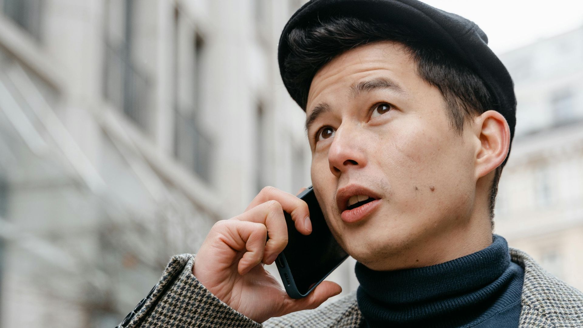 Stylish man in a city talking on a smartphone outdoors, wearing a coat and hat.