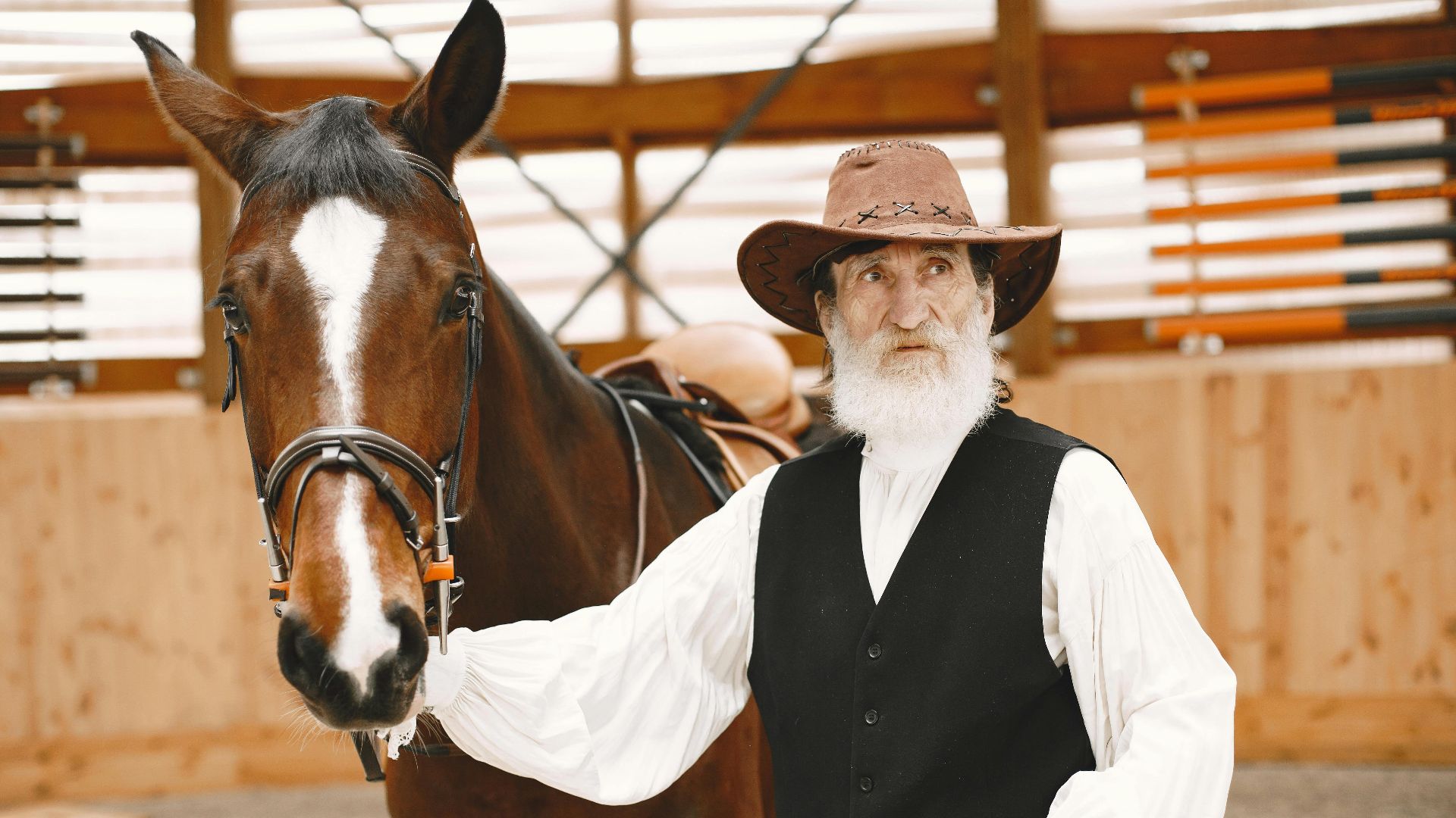 Elderly man in cowboy hat beside a brown horse, standing in a rustic barn.
