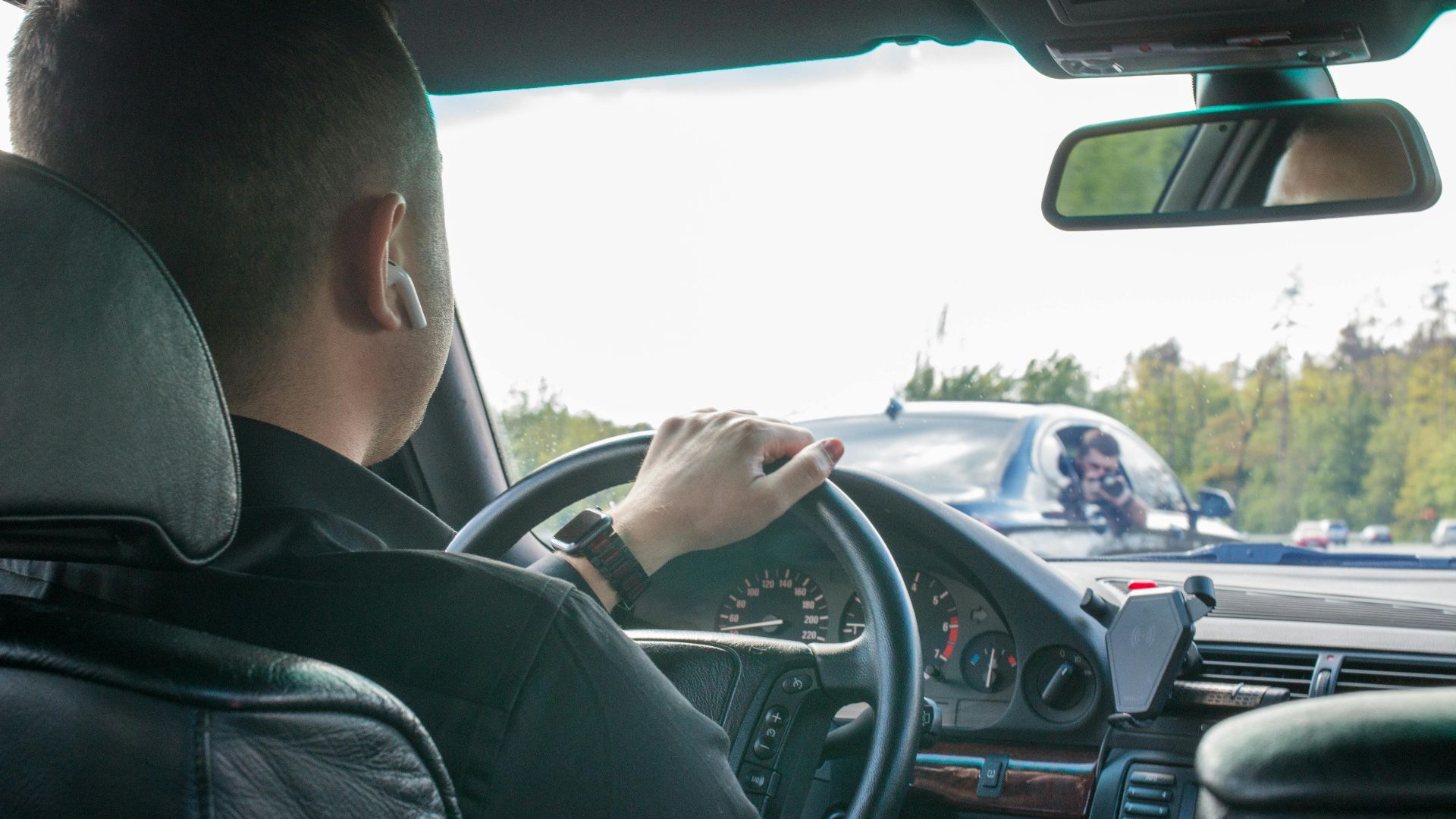 A man is driving a car seen from the back seat, focusing on the road ahead.