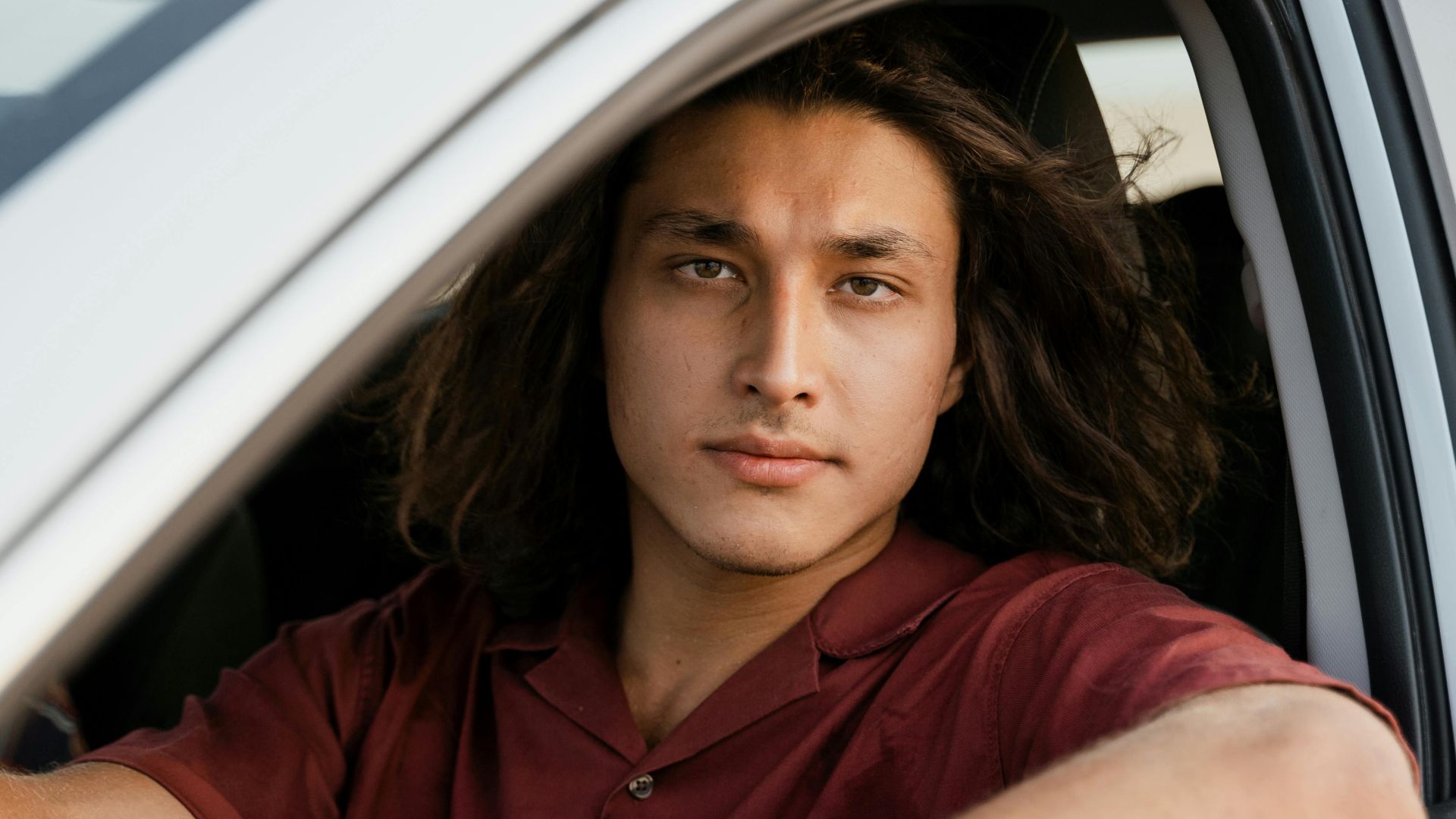 Portrait of a young man in a red shirt, sitting inside a car, ready to drive.