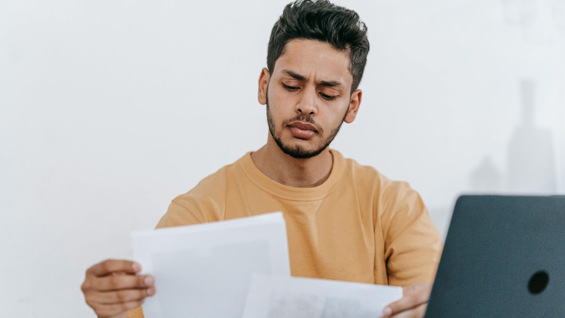 Attentive young bearded Hispanic male remote worker examining papers with results of project while working with laptop in home office