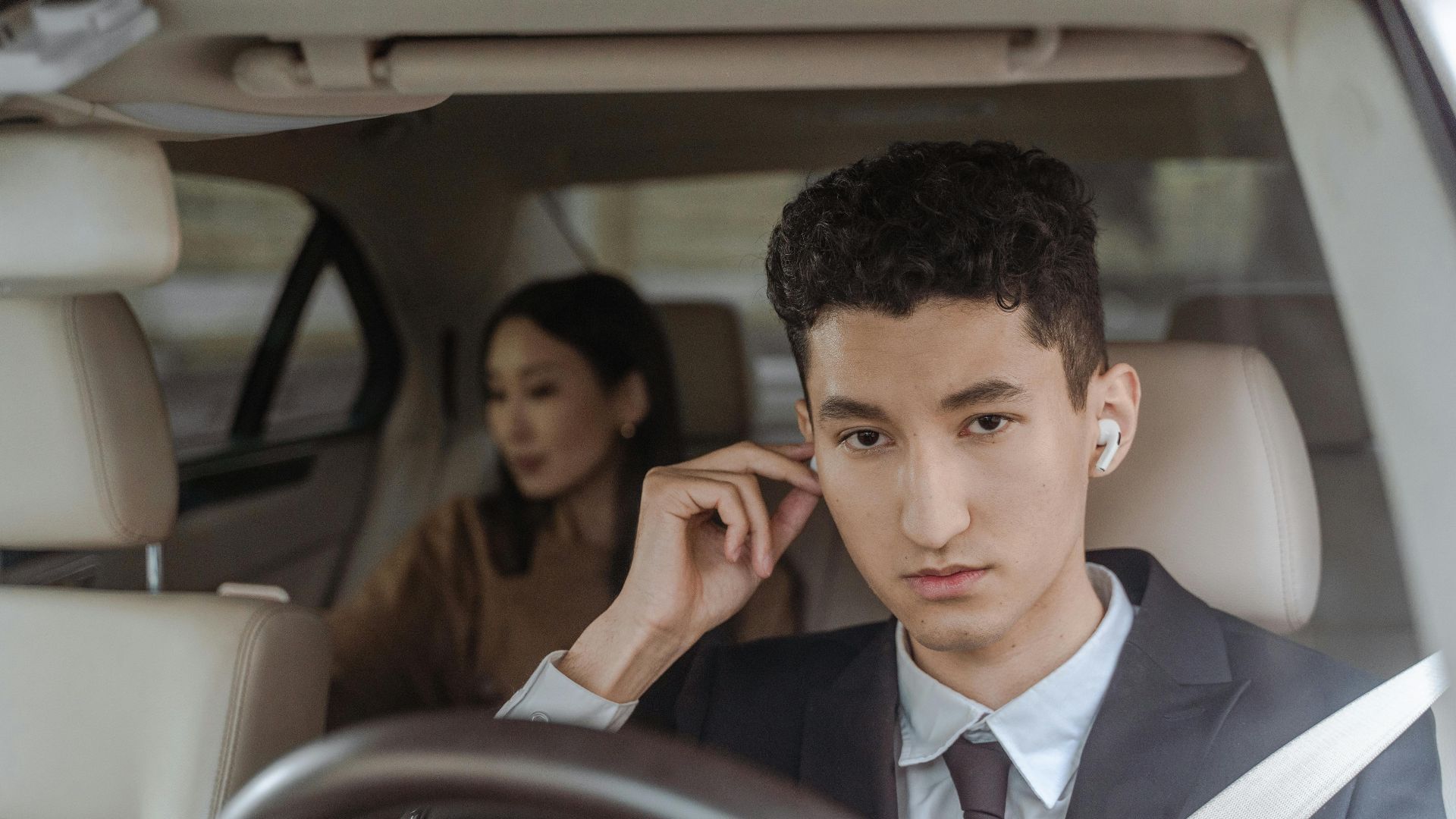 A professional driver in a suit with AirPods driving a female passenger in a car.