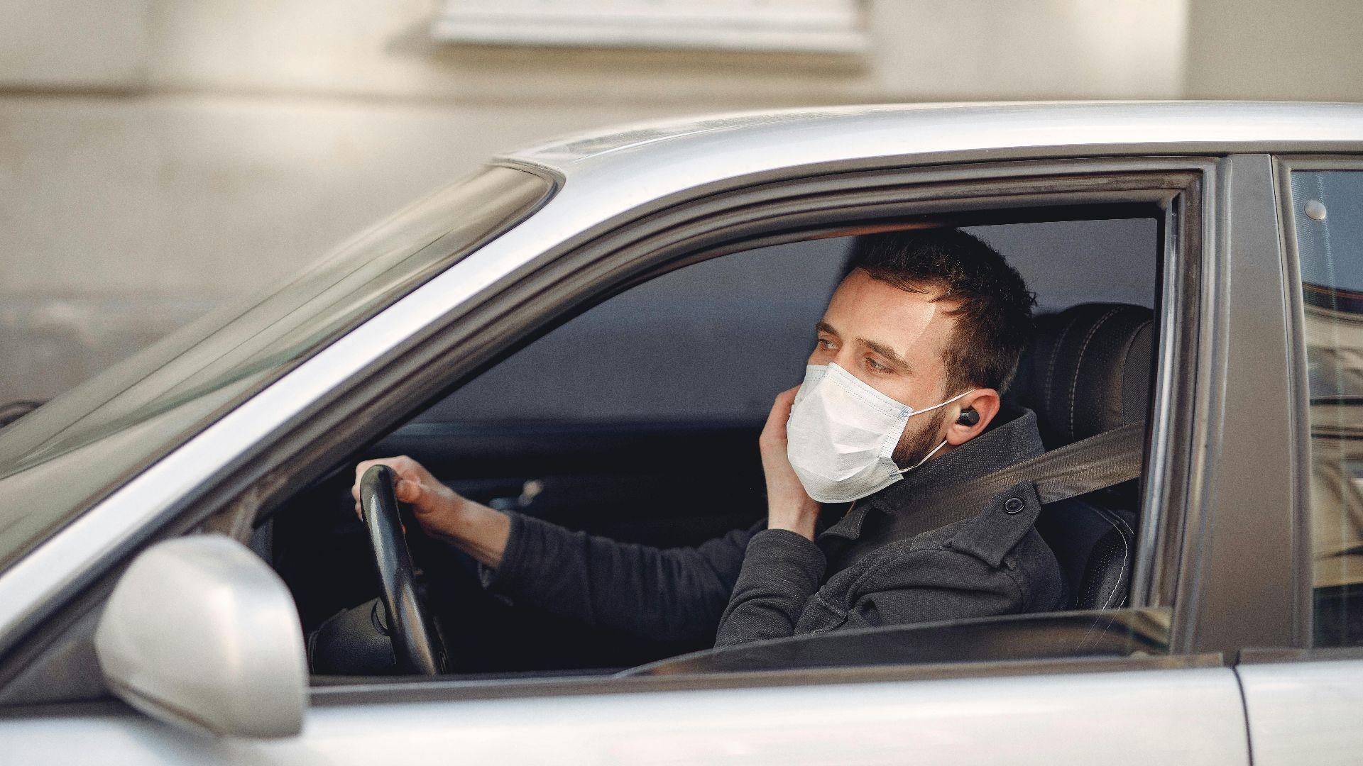 Serious bearded male driver wearing warm jacket and protective facial mask using wireless earphones sitting in auto with opened window during coronavirus pandemic