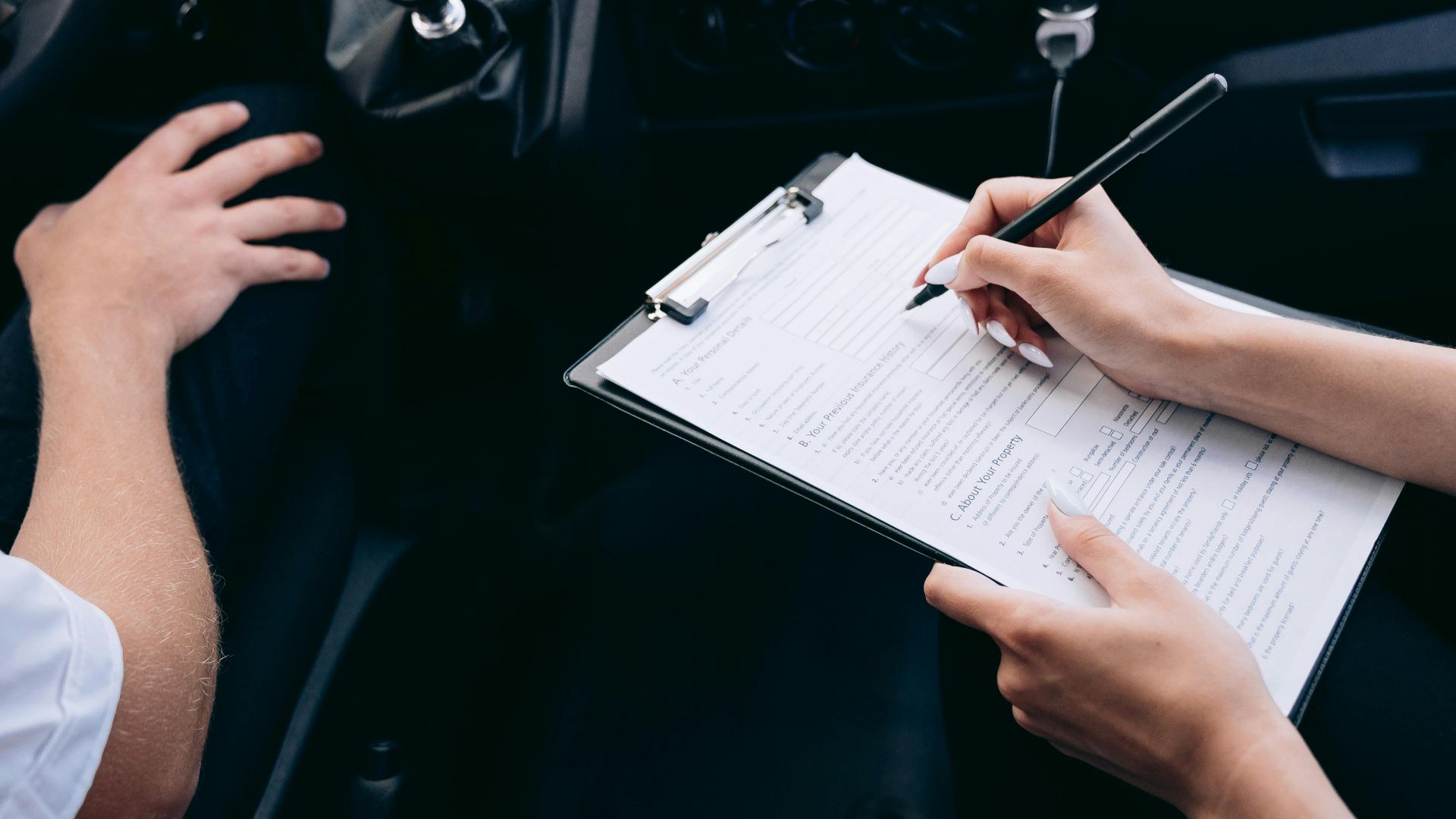 Close-up of a person writing on a clipboard inside a car, showing hands and a gear shift.