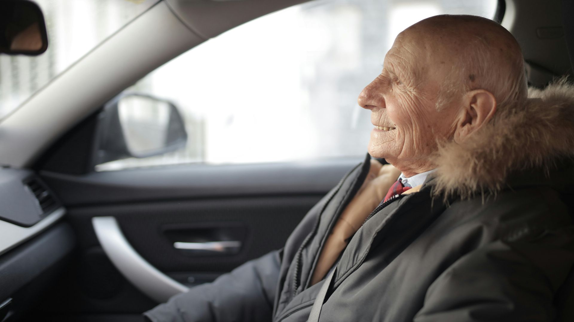 Side view of content elderly male in suit and outerwear sitting in front seat of contemporary automobile and looking away