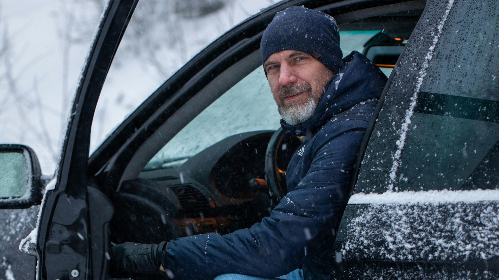 A man in winter attire sits in a car doorway amid falling snow, capturing a serene winter moment.