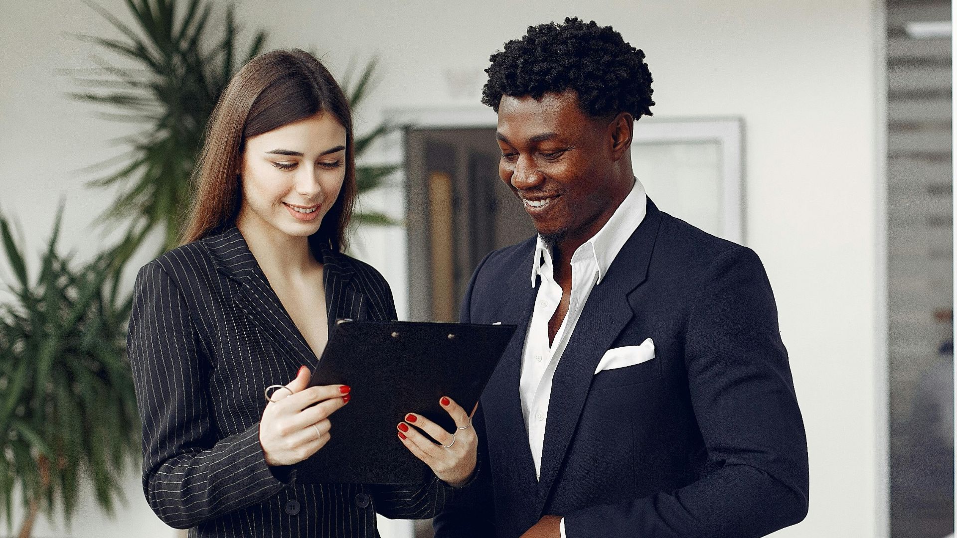 Cheerful multiethnic stylish female agent and smiling customer in formal suit discussing contract details while standing in modern car showroom in daylight