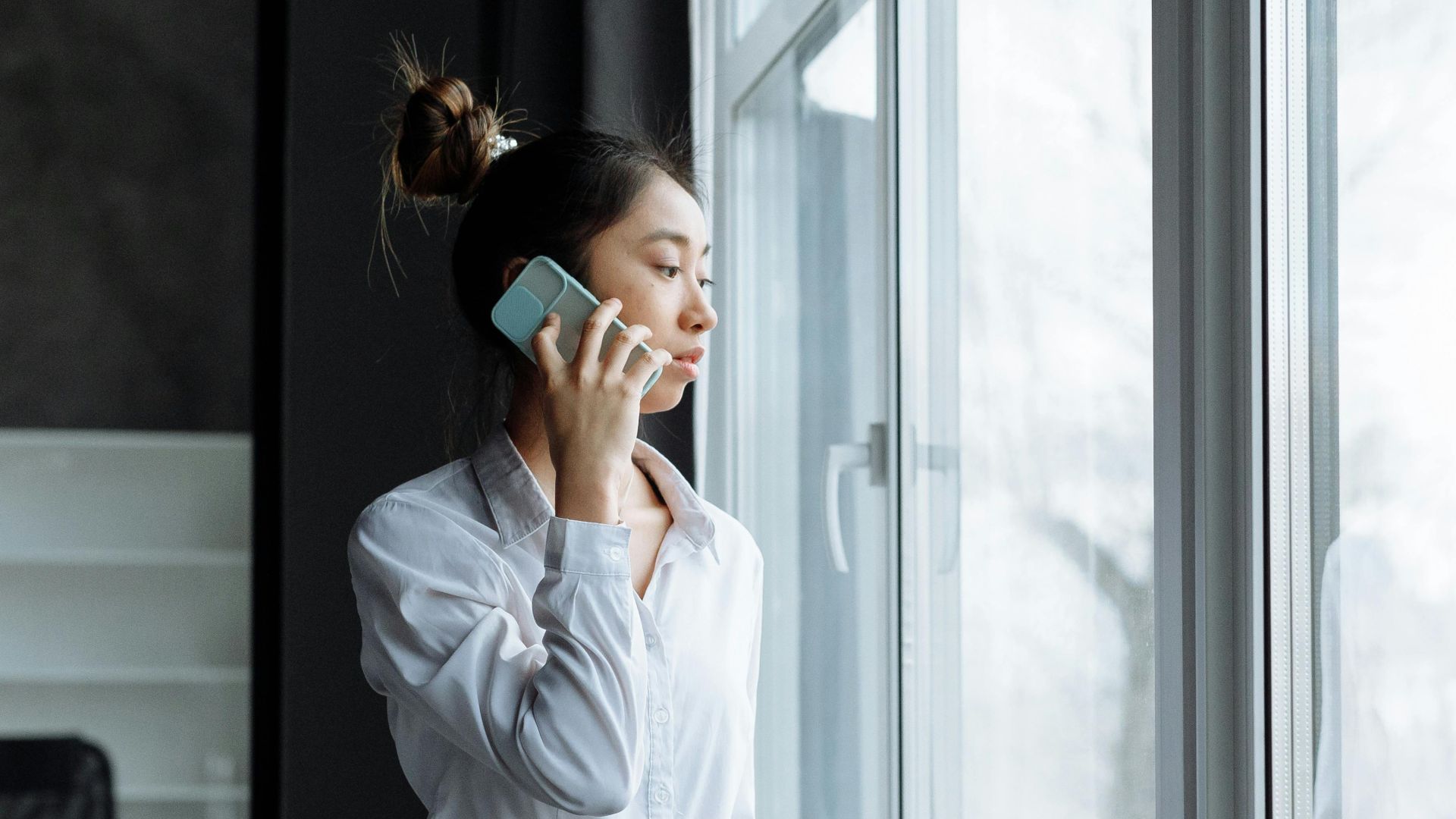 Asian woman in office clothes making a phone call while gazing outside a large window.