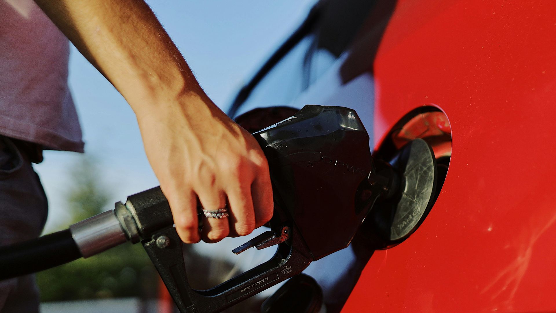 Close-up of a person refueling a red car at an outdoor gas station during the day.