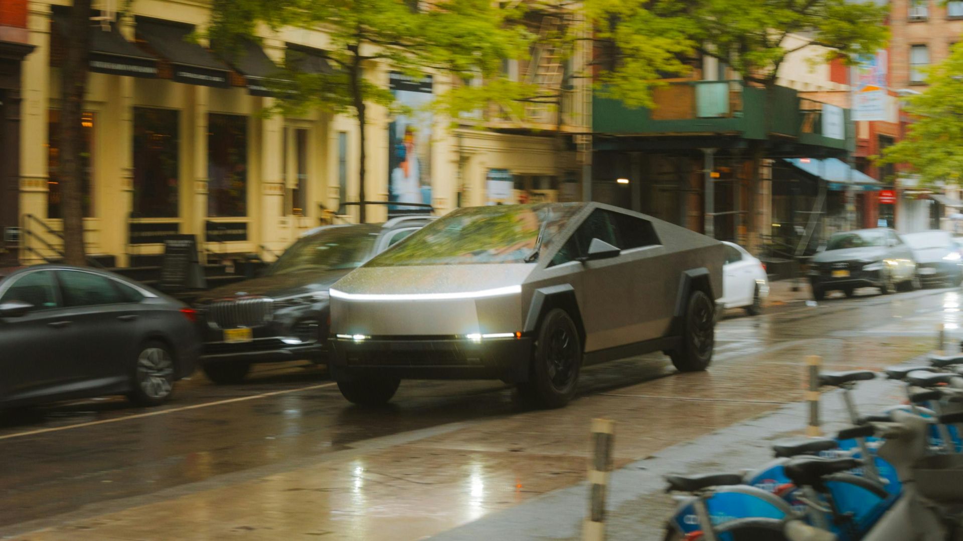 A sleek Cybertruck driving on a rainy city street lined with bicycles and buildings.