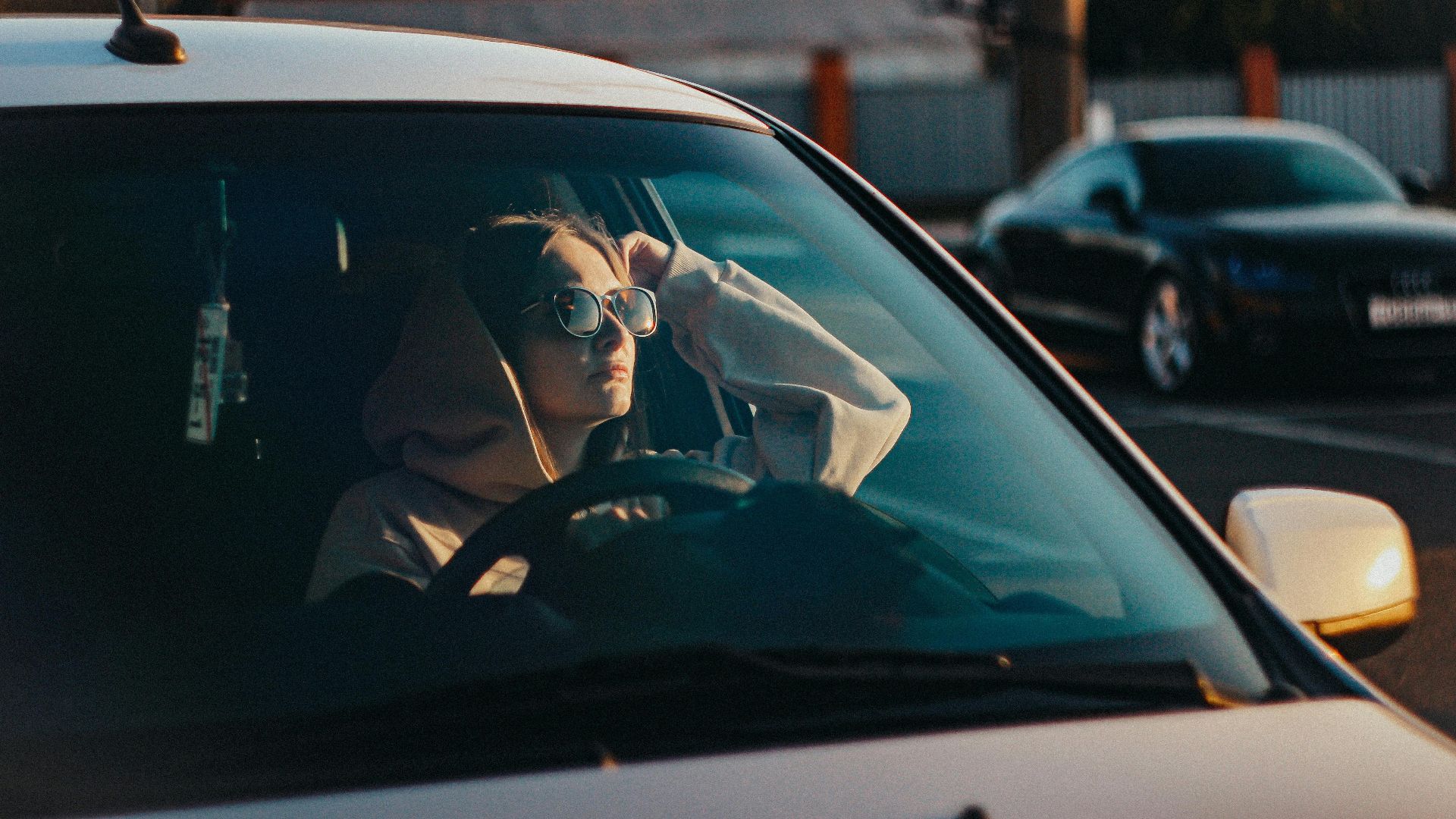 Woman in hoodie and sunglasses relaxing in a parked car at sunset.