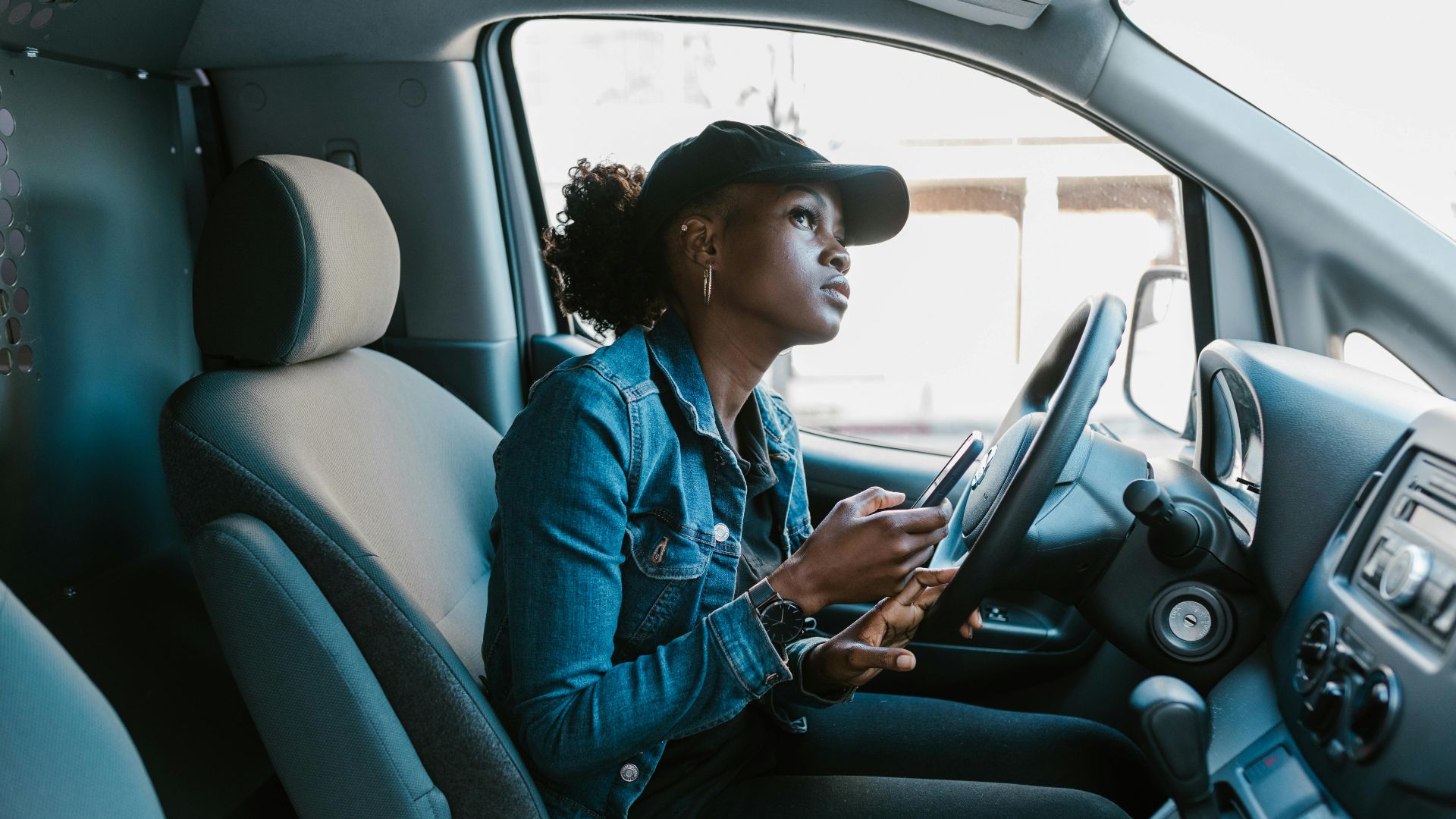A woman in a denim jacket focuses on her smartphone while sitting in a car driver's seat.