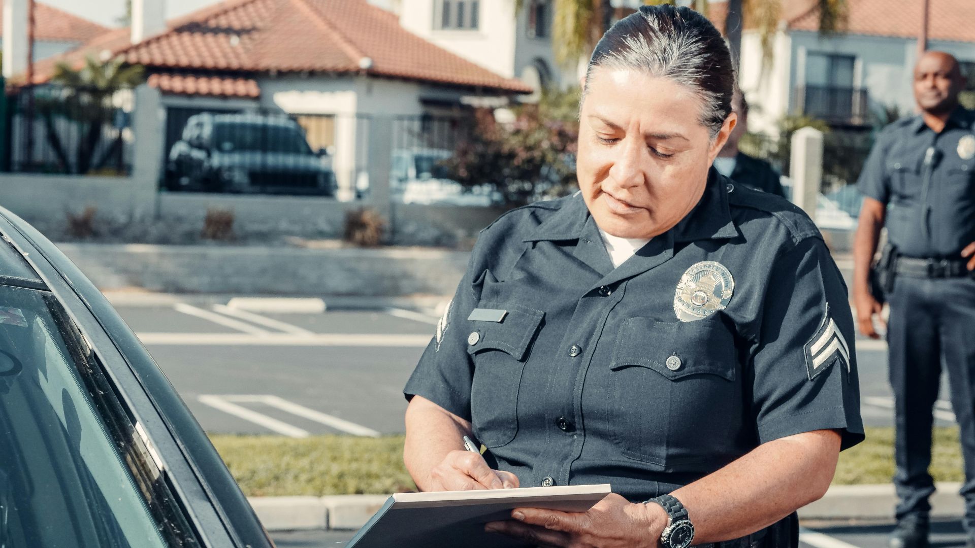 Police officer writing a ticket to a car in a sunny parking lot with a colleague in the background.