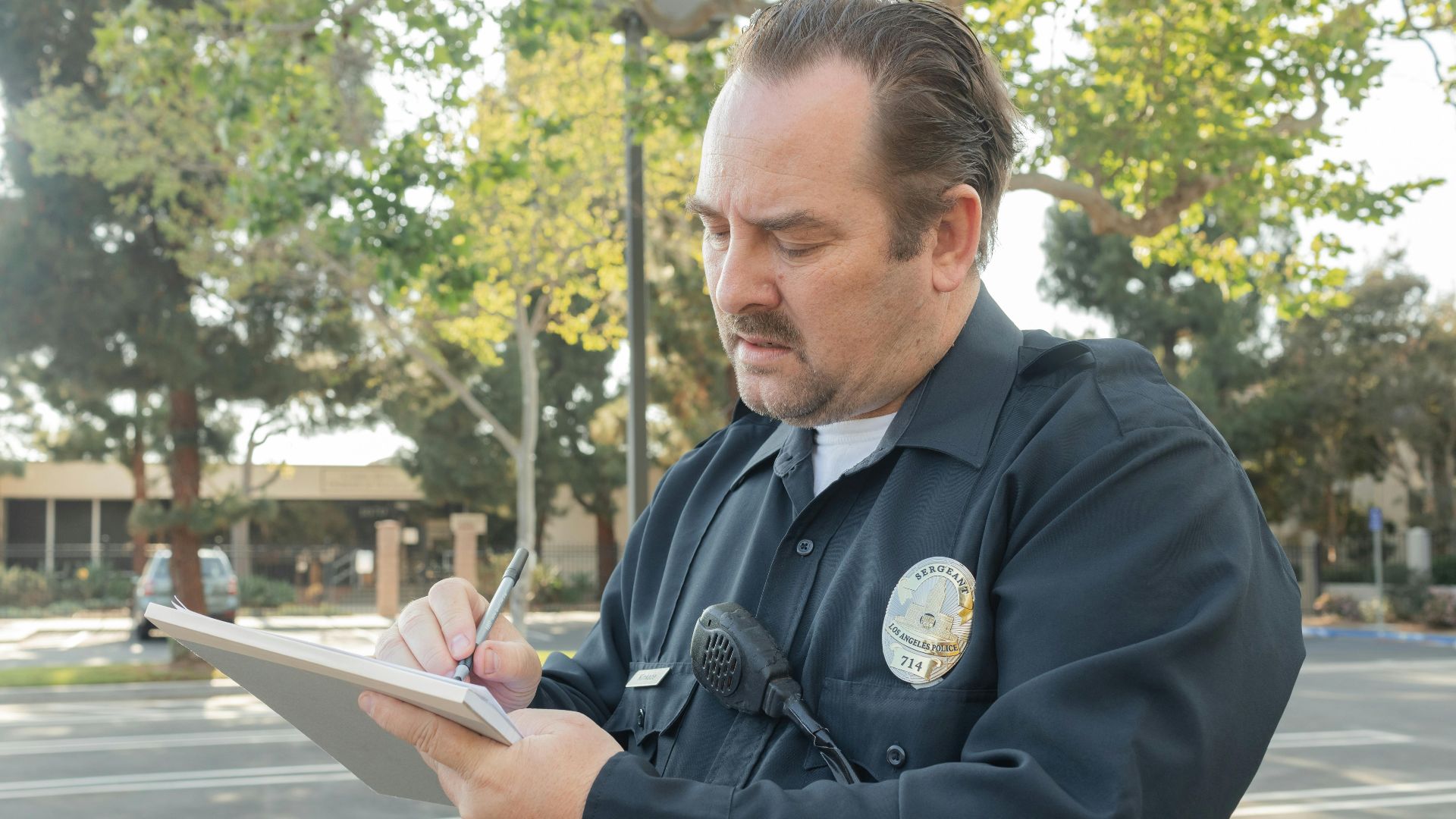 A police officer outdoors writing a report on a notepad. Trees in the background.