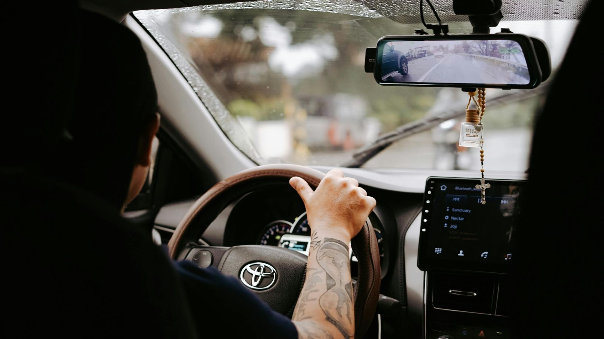 A driver's tattooed arm on the steering wheel inside a Toyota car during the day.