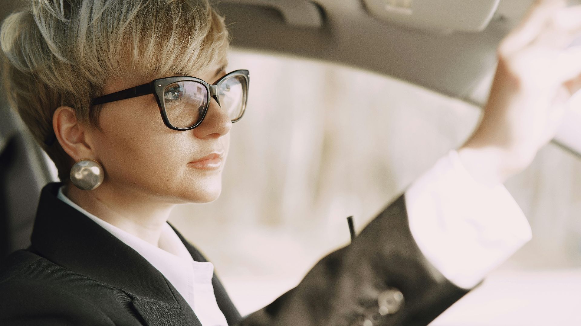Side view of crop female driver in stylish formal outfit and eyeglasses with takeaway coffee looking at rear mirror of modern car