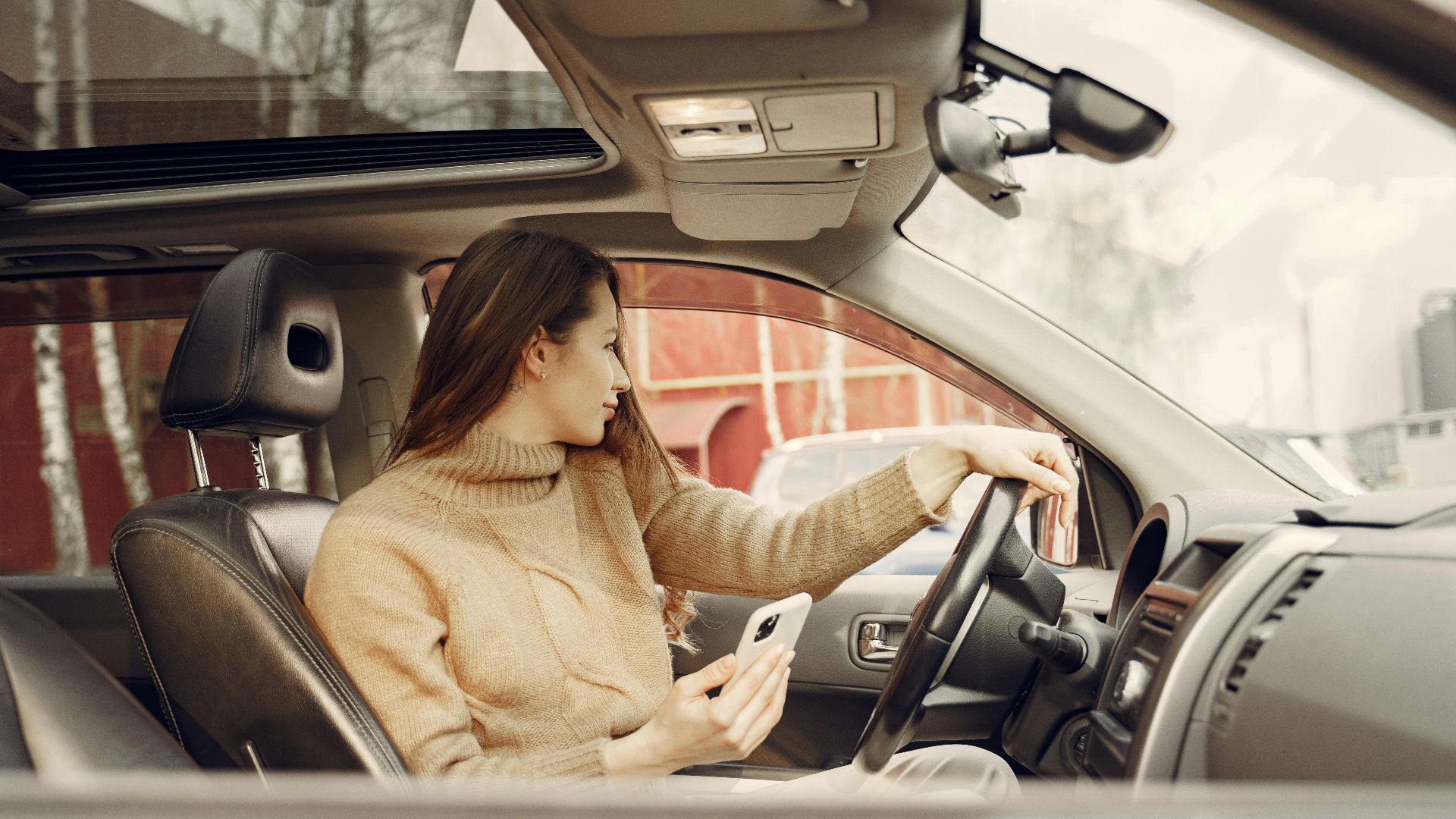 From window side view of adult woman in warm clothes sitting at wheel of modern car waiting for friend and using smartphone