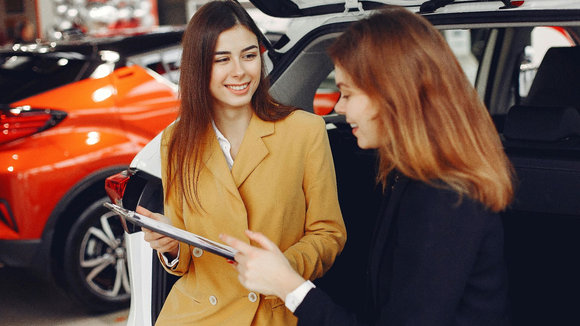 Positive smiling agent in trendy formal light brown suit discussing with happy excited female customer details of contract while leaning on car in showroom