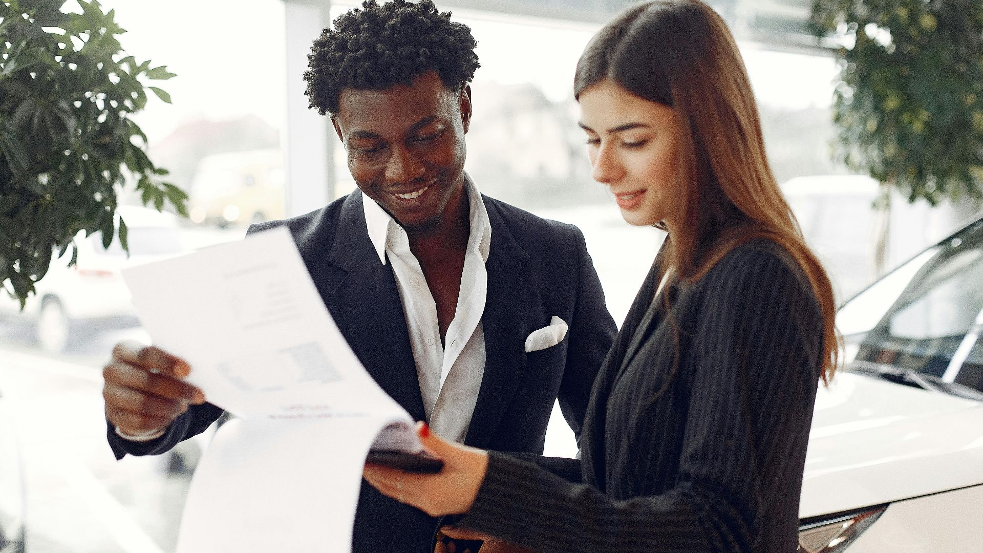 Cheerful smiling multiethnic businessman in classy suit and professional elegant female consultant standing close and reading contract details in car showroom