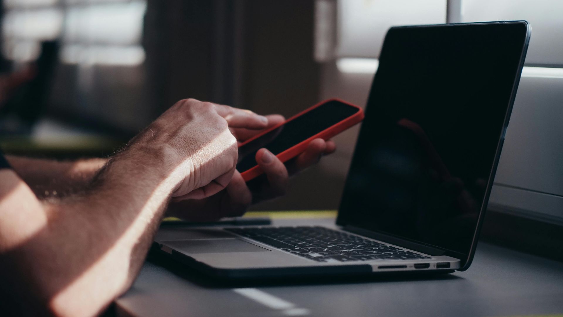 A person holding a smartphone near a laptop in an indoor setting, depicting modern technology use.