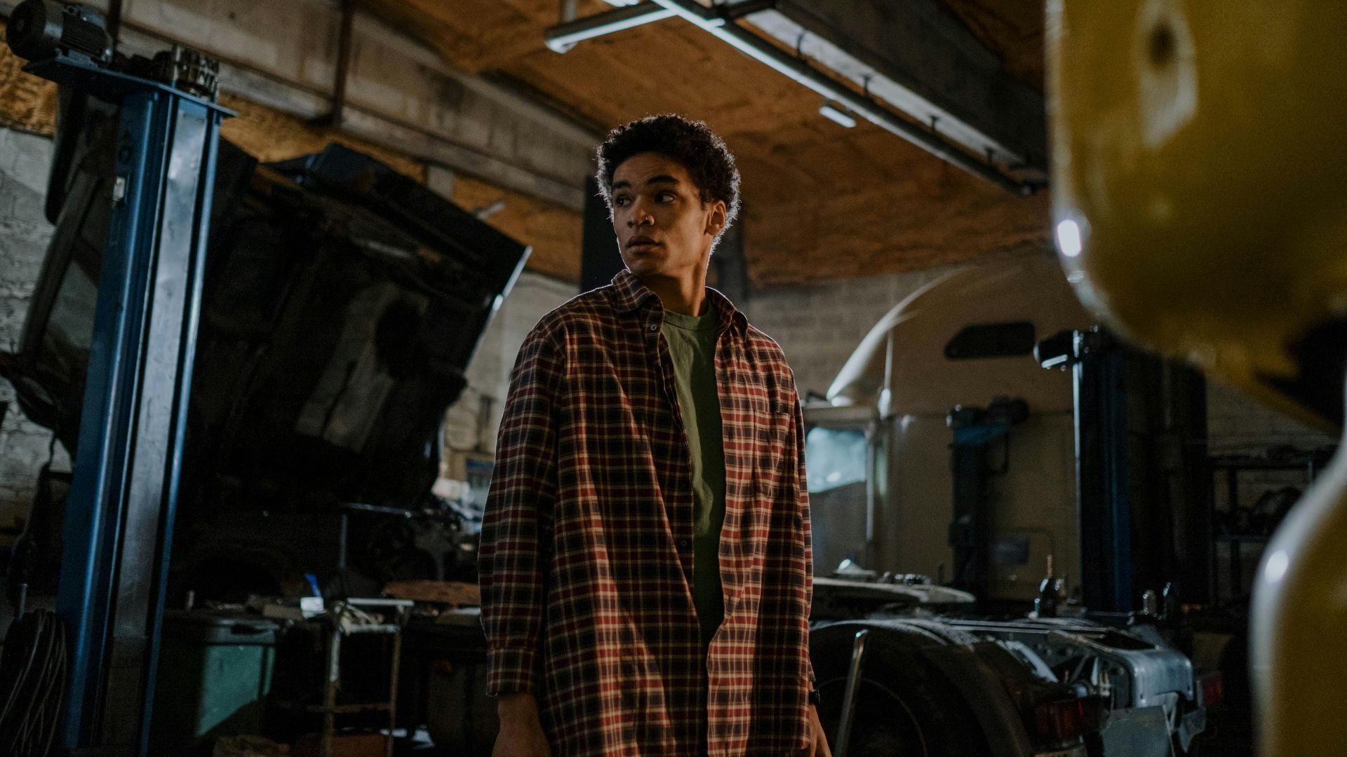 A young mechanic in a plaid shirt stands in a dimly lit auto workshop, deep in thought.