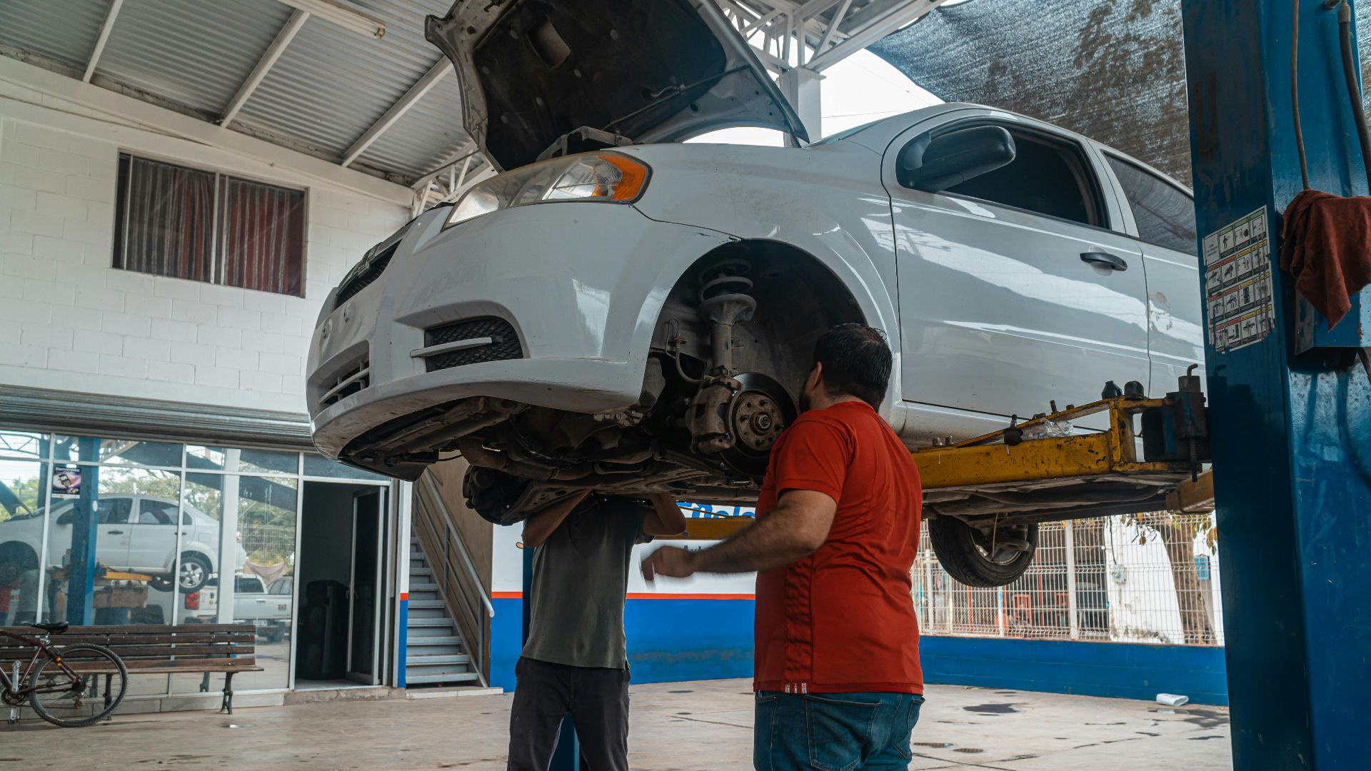 Two mechanics inspecting a white car on a lift inside a garage workshop.