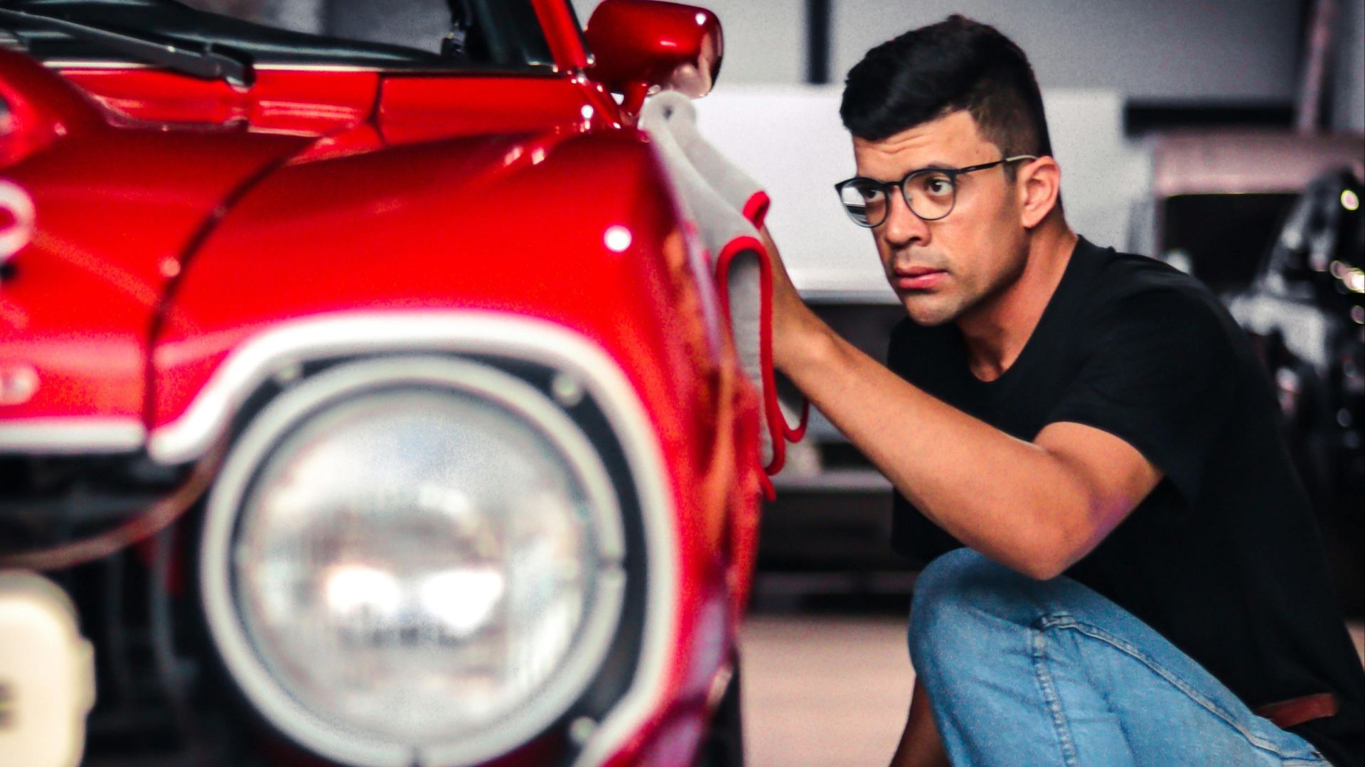 A man kneels to inspect a classic red car in a garage setting, emphasizing detail and care.
