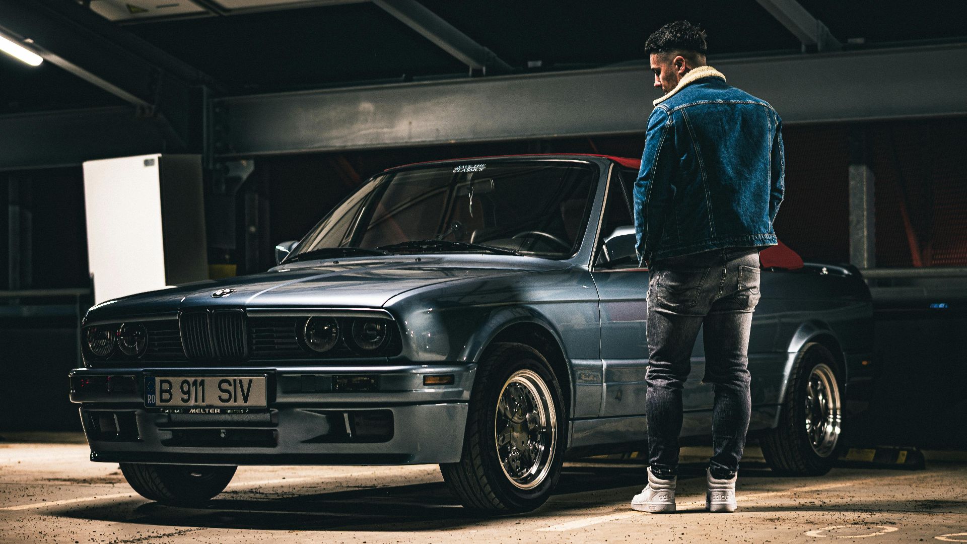 A man in a denim jacket stands beside a vintage blue car in an indoor parking lot.