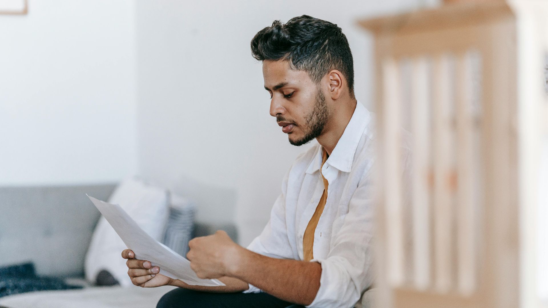 Concentrated young ethnic male wearing casual outfit reading documents and sitting on couch in modern living room