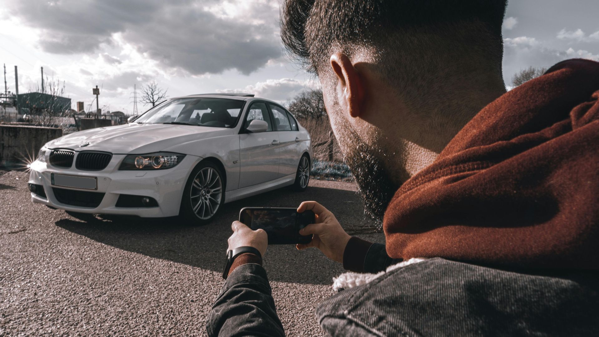 A man takes a picture of a white luxury car using a smartphone outdoors under a cloudy sky.