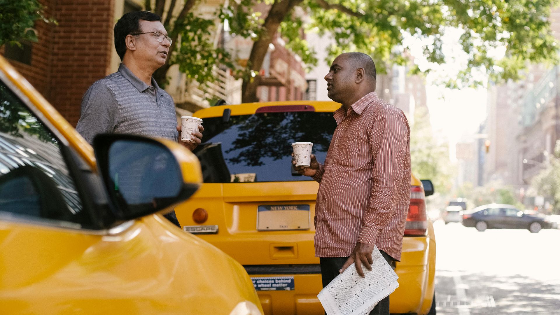 Side view of multiracial male coworkers with newspaper and takeaway coffee speaking near taxi autos while looking at each other in town