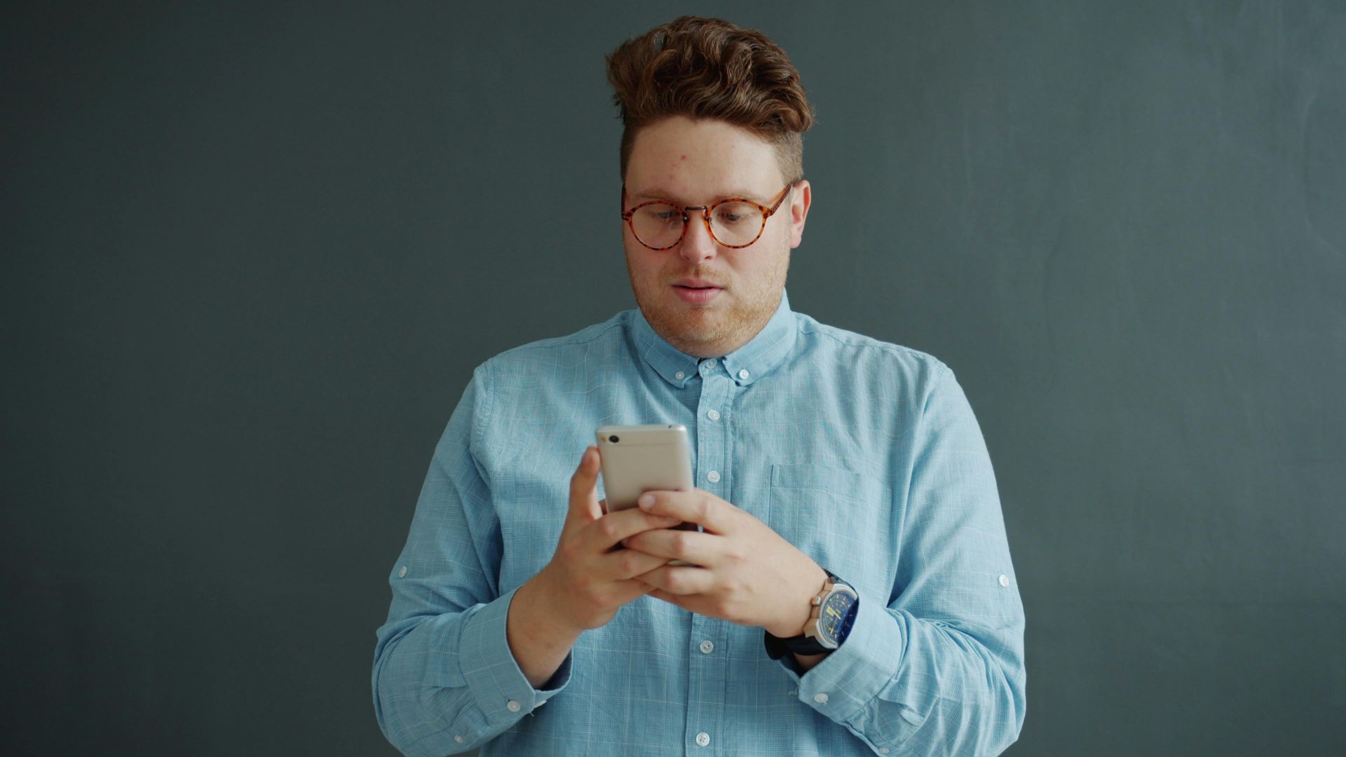 Young adult wearing glasses using a smartphone indoors with a neutral background.