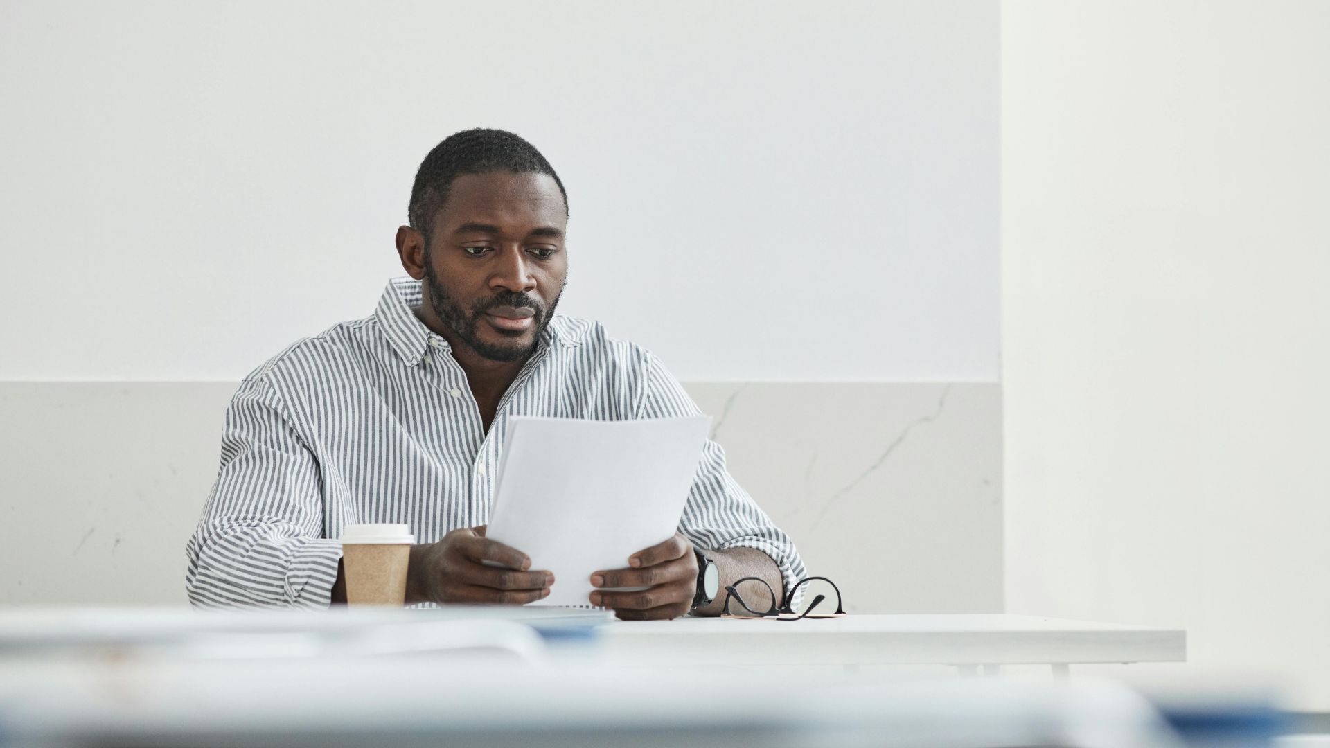African American man sitting indoors, reading papers with a coffee cup nearby.